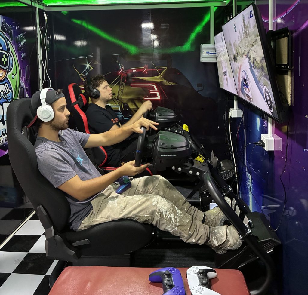 Two men competing in professional racing simulators with Thrustmaster steering wheels and racing cockpits inside a mobile gaming trailer with green LED lighting for corporate team building in Sydney.